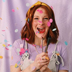 Lydia Packham with a wide mouth smile holding calligraphy dip pens with confetti falling in foreground